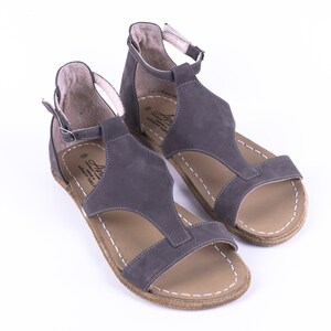 Puede incluir: Un par de sandalias de cuero gris con correas de tobillo. Las sandalias tienen un dise&ntilde;o de correa en T y una suela de cuero marr&oacute;n.