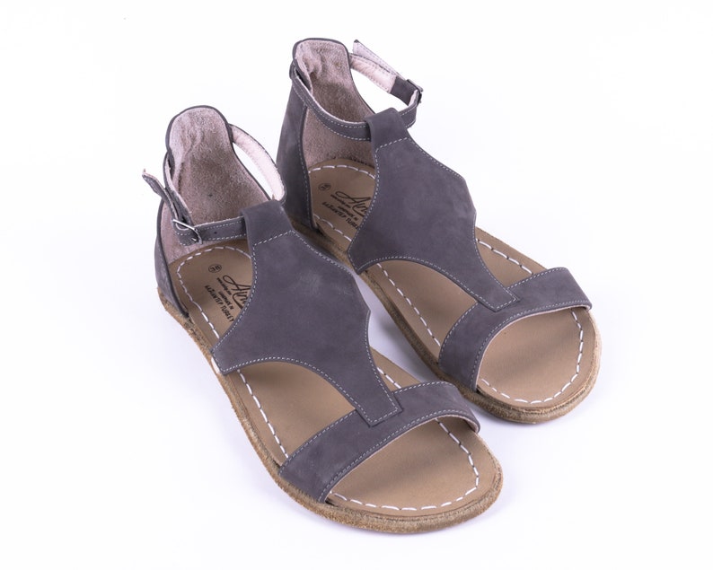Puede incluir: Un par de sandalias de cuero gris con correas de tobillo. Las sandalias tienen un dise&ntilde;o de correa en T y una suela de cuero marr&oacute;n.
