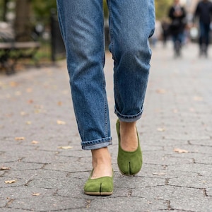 以下が含まれることがあります： 青いジーンズと緑色の足袋のような靴を履いた人がレンガの小道を歩いています。靴はくすんだ緑色で、親指と他の指を分けるユニークなデザインです。