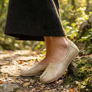 Op de afbeelding: Tan leren split-teen balletschoenen. De schoenen hebben een ronde neus en een lage hak. De schoenen worden buiten op een stenen pad gedragen. De schoenen hebben een neutrale kleur.