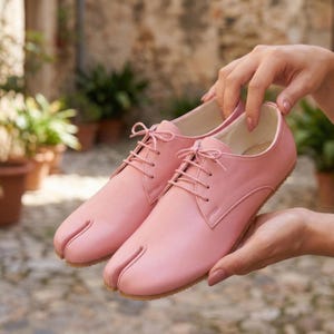 Op de afbeelding: Een paar roze schoenen met split-teen en veters. De schoenen zijn gemaakt van glad materiaal en hebben een lichtbruine zool. Het ontwerp heeft een split-teen. De schoenen worden door handen vastgehouden.