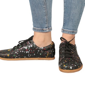 Op de afbeelding: Zwarte canvas lage sneakers met een kleurrijk verfspatten ontwerp. De schoenen hebben zwarte veters en een bruine rubberen zool.