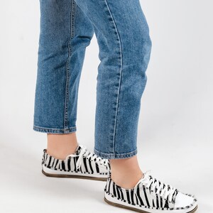 May include: A pair of white and black zebra print canvas sneakers with white laces. The sneakers are worn by a person wearing blue jeans.