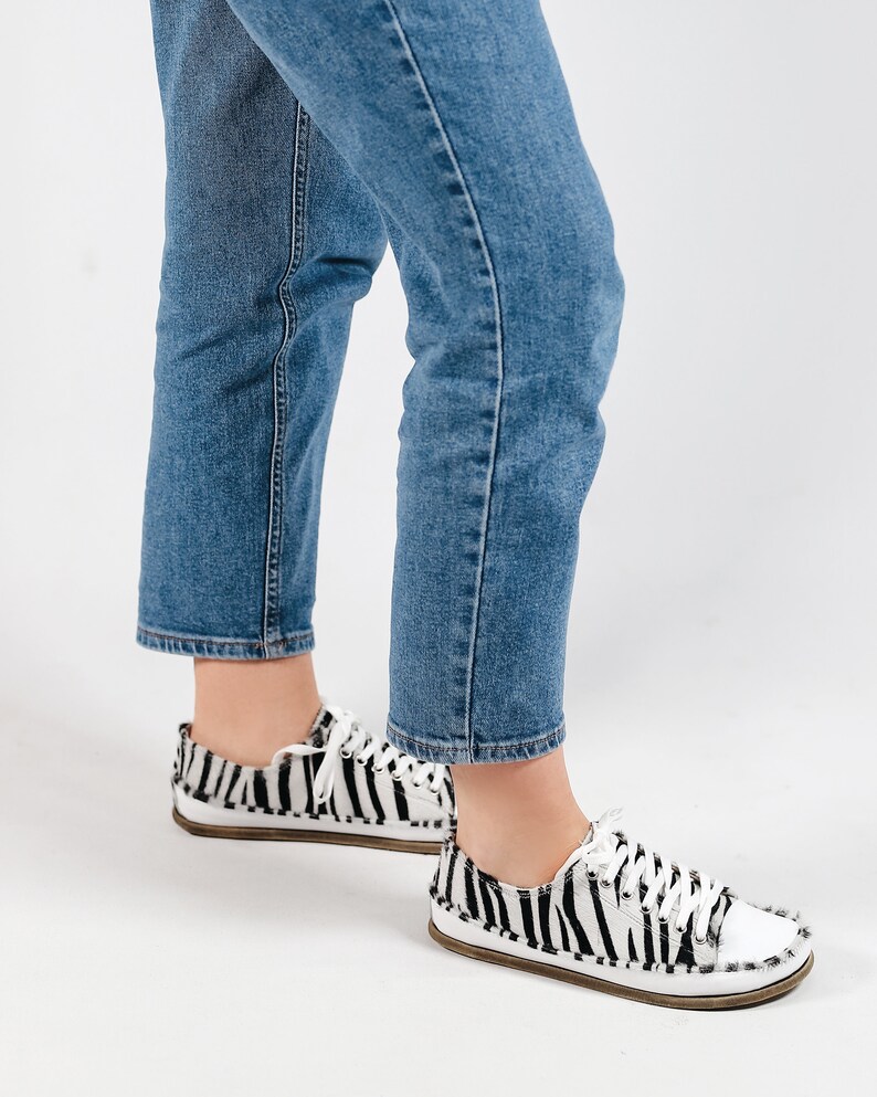 May include: A pair of white and black zebra print canvas sneakers with white laces. The sneakers are worn by a person wearing blue jeans.