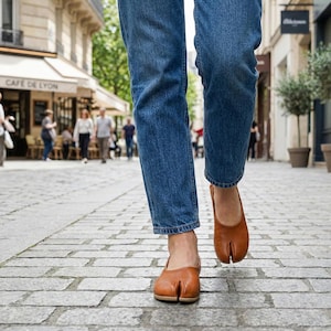 Op de afbeelding: Een persoon loopt op een geplaveide straat met een blauwe jeans en bruine leren schoenen met split-teen. De schoenen hebben een uniek ontwerp met een apart compartiment voor de grote teen.