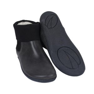 Puede incluir: Un par de botines negros con un puño de punto negro en contraste. Una bota está en posición vertical, mientras que la otra se muestra con la suela hacia arriba. Las suelas son de color gris oscuro con un diseño texturizado. Las botas parecen ser de cuero.