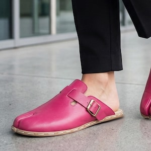 May include: A pair of bright pink leather clogs with a buckle closure. The clogs have a toe separator and a light brown sole with white stitching. The background shows black pants and a gray surface.