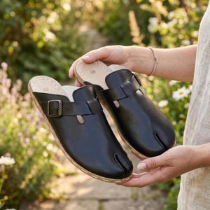 May include: A pair of black leather clogs with a split toe design. The clogs have a buckle closure and a light brown sole. The shoes are held up in front of a blurred background of greenery.