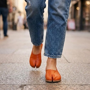 May include: Orange leather split-toe shoes, also known as Tabi shoes, are worn with blue denim jeans. The shoes have a unique design with a separation for the big toe. The person is walking on a stone surface.