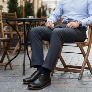 May include: Black leather dress shoes with brown soles. The shoes are laced up and worn with dark gray dress pants and navy blue socks. The person is sitting on a wooden chair.