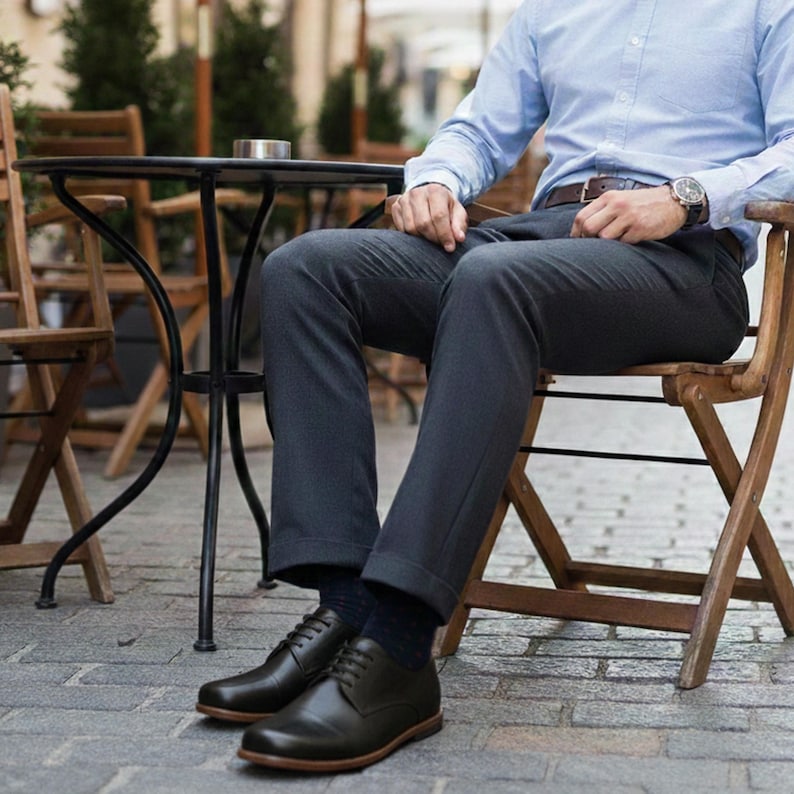 May include: Black leather dress shoes with brown soles. The shoes are laced up and worn with dark gray dress pants and navy blue socks. The person is sitting on a wooden chair.
