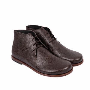 May include: A pair of brown leather chukka boots with a textured finish. The boots have a lace-up design and a rounded toe. The soles are a darker brown color. The boots are set against a white background.