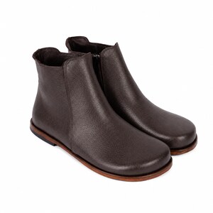 May include: A pair of dark brown ankle boots featuring a rounded toe, a smooth textured upper, and a side zipper closure. The boots are constructed with a flat, layered brown sole and a low heel, providing a clean and simple silhouette.