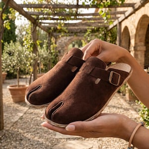 May include: A pair of brown suede clogs with a split-toe design. The clogs have a strap with a gold-colored buckle. The shoes are held in a person's hands, with a blurred outdoor background.