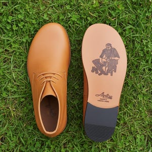 Puede incluir: Un par de zapatos de vestir de cuero color canela. Uno se ve desde arriba, el otro revela la suela con un gr&aacute;fico de una persona y el nombre "Anton". Los zapatos est&aacute;n sobre hierba verde.