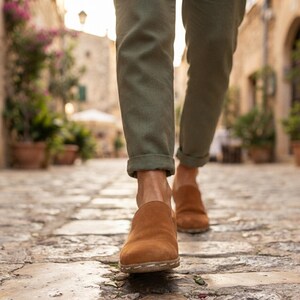 May include: A pair of tan suede slip-on shoes with visible light-colored stitching along the sole, worn with olive green cuffed trousers. The footwear features a rounded toe and a low-profile design, captured while walking on a stone path.