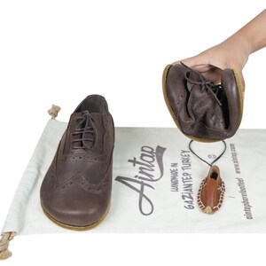May include: A pair of dark brown leather oxford shoes with brogue detailing and thin laces. One shoe is held and folded to demonstrate flexibility, while the other sits on a white fabric bag printed with the text Aintap Handmade in Gaziantep Turkey.