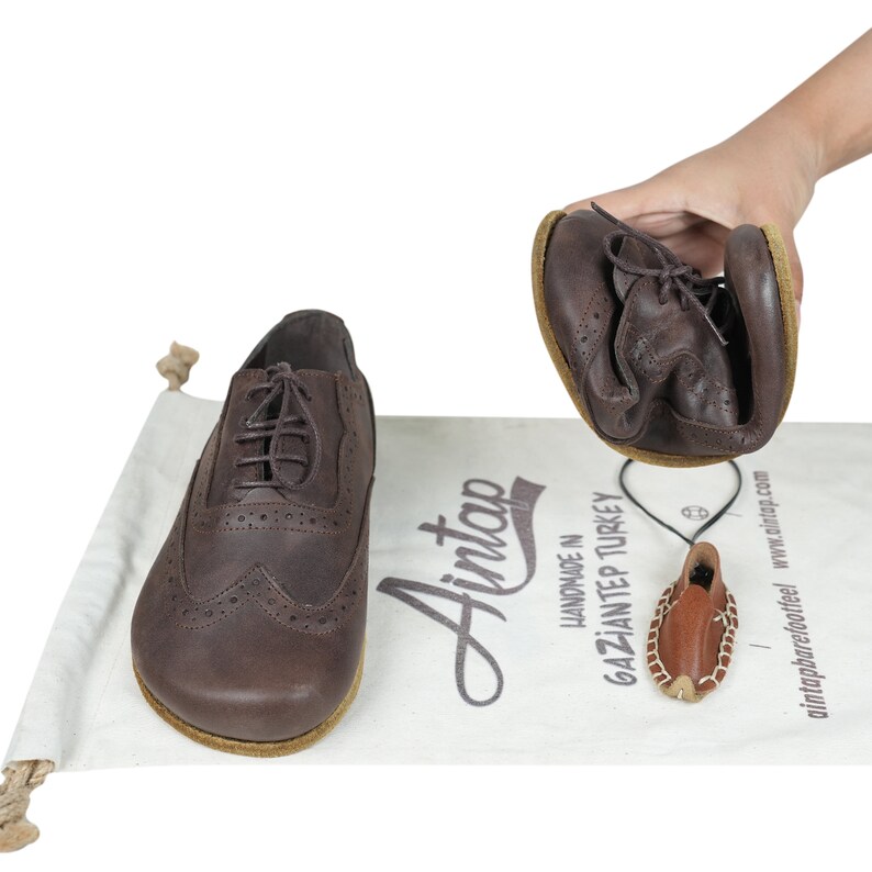 May include: A pair of dark brown leather oxford shoes with brogue detailing and thin laces. One shoe is held and folded to demonstrate flexibility, while the other sits on a white fabric bag printed with the text Aintap Handmade in Gaziantep Turkey.