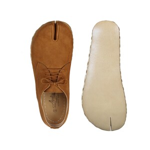 Puede incluir: Un par de zapatos marrones de ante con puntera dividida, cordones y suela de color claro. Los zapatos tienen un diseño único con una división entre el dedo gordo y los otros dedos. La suela es de color beige claro con un borde cosido.