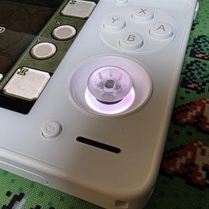Puede incluir: Consola de juegos portátil blanca con una pantalla que muestra una interfaz de juego. La pantalla muestra las palabras "No Map" e iconos de juego. La consola tiene un pad direccional, botones y un joystick con una luz brillante. El fondo es un patrón pixelado verde.