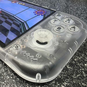May include: A handheld gaming console with a transparent plastic shell, featuring a color screen displaying a video game with a checkered floor. The device includes a white analog joystick, four circular buttons labeled A, B, X, and Y, and a power button.