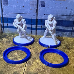 May include: Two white plastic miniature figures of soldiers wearing helmets and holding guns, standing on white bases with blue rings around them.