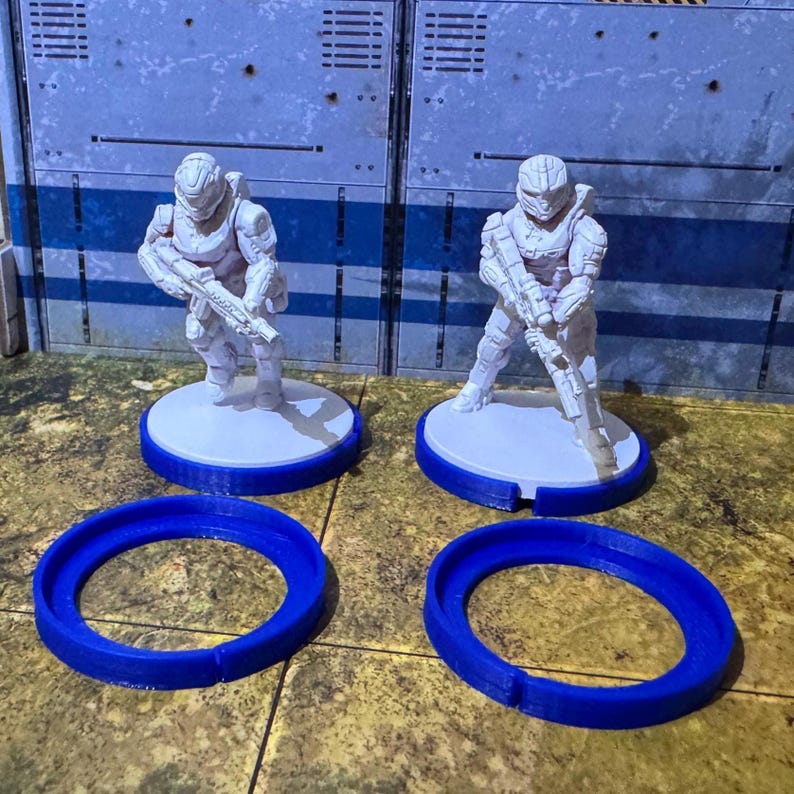 May include: Two white plastic miniature figures of soldiers wearing helmets and holding guns, standing on white bases with blue rings around them.