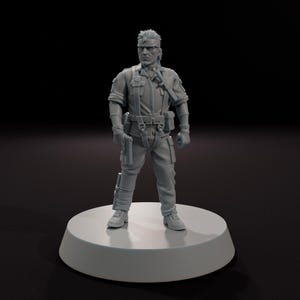以下が含まれることがあります： 白い円形の台座に立っている、軍事スタイルの制服を着た男性の灰色のフィギュア。フィギュアは銃を持っており、頭に包帯をしています。制服には、タクティカルベスト、手袋、ブーツが含まれています。背景は黒です。