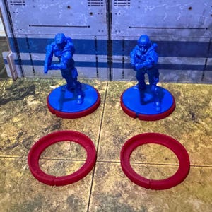 May include: Two blue plastic miniature figures standing on blue bases with red rings. The figures are wearing helmets and holding weapons. Two additional red rings are on the surface.
