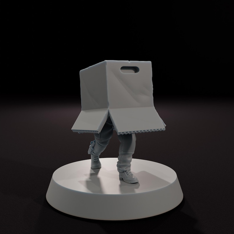 以下が含まれることがあります： 頭に箱をかぶった人物の灰色の置物。箱のフラップは開いています。人物は灰色のズボンとスニーカーを履き、白い円形の台座の上に立っています。