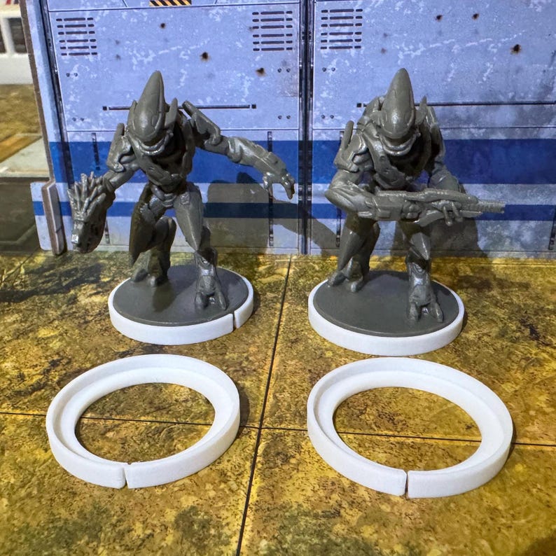 May include: Two grey plastic action figures of alien warriors standing on white circular bases. The figures are posed in a dynamic stance, one holding a weapon and the other with its arm raised. The figures are on a textured surface.