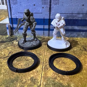 May include: Two miniature figures, one grey and one white, stand on circular bases. The bases are on top of black plastic rings. The figures are wearing futuristic armour and helmets.