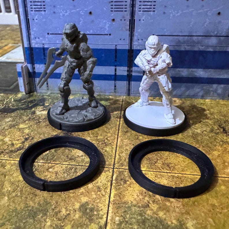 May include: Two miniature figures, one grey and one white, stand on circular bases. The bases are on top of black plastic rings. The figures are wearing futuristic armour and helmets.