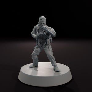 以下が含まれることがあります： 銃とナイフを持った戦闘服姿の兵士の灰色のフィギュア。フィギュアは、暗い背景に対して、白い円形の台座の上に立っています。兵士はブーツ、ズボン、ベスト、長袖シャツを着ています。