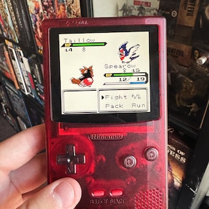 Może przedstawiać: Czerwona przenośna konsola do gier Game Boy Color z przezroczystą plastikową obudową. Na ekranie wyświetlana jest walka Pokémonów między Taillowem a Spearowem. Tekst na ekranie brzmi "Taillow HP 14/18", "Spearow ♀15 HP 12/19", "Fight Run Pack Run". Konsola ma logo Nintendo i przyciski.