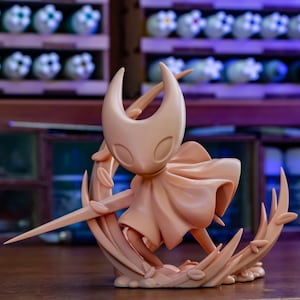 May include: A light peach-colored figurine of a character with a horned head, cape, and a long, thin weapon. The figure is posed within a crescent-shaped base with leaf details. The background features shelves with decorative items.