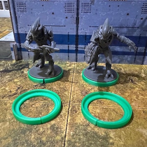May include: Two grey plastic miniature figures of alien warriors standing on green plastic bases. The figures are holding weapons and are posed in a fighting stance.