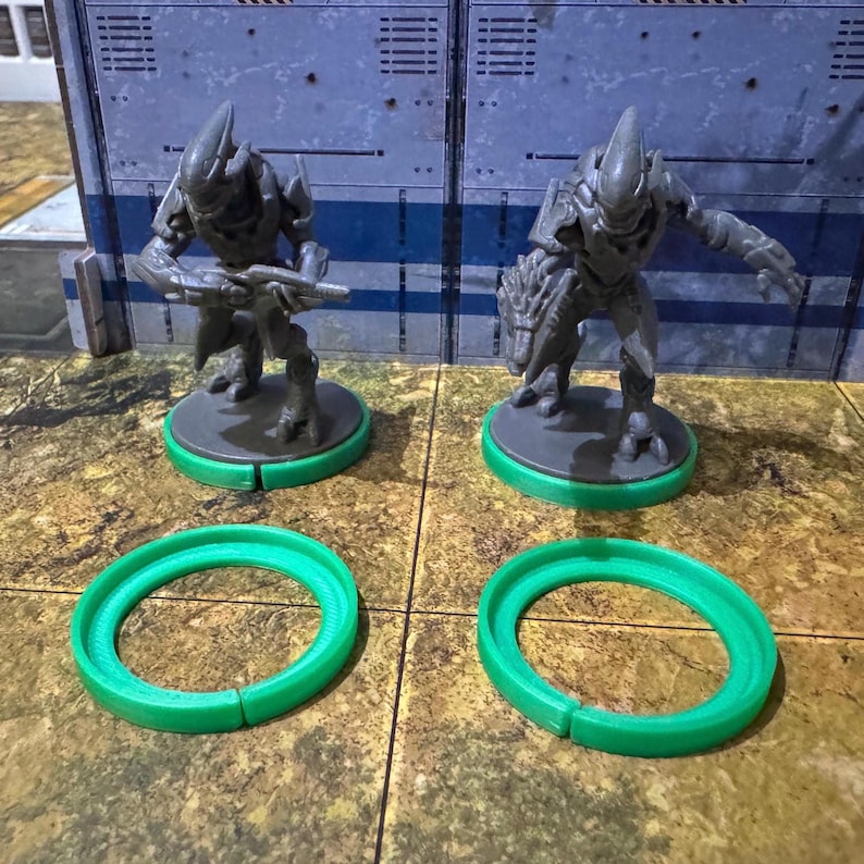 May include: Two grey plastic miniature figures of alien warriors standing on green plastic bases. The figures are holding weapons and are posed in a fighting stance.