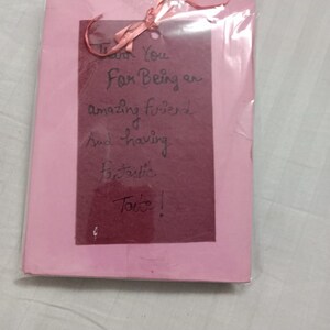 Puede incluir: Una tarjeta de felicitaci&oacute;n rosa con un inserto rectangular rojo oscuro, envuelta en pl&aacute;stico transparente. Una cinta rosa est&aacute; atada en la parte superior. El texto manuscrito en el inserto dice: "Thank You For Being an amazing friend And having fantastic Taste!"
