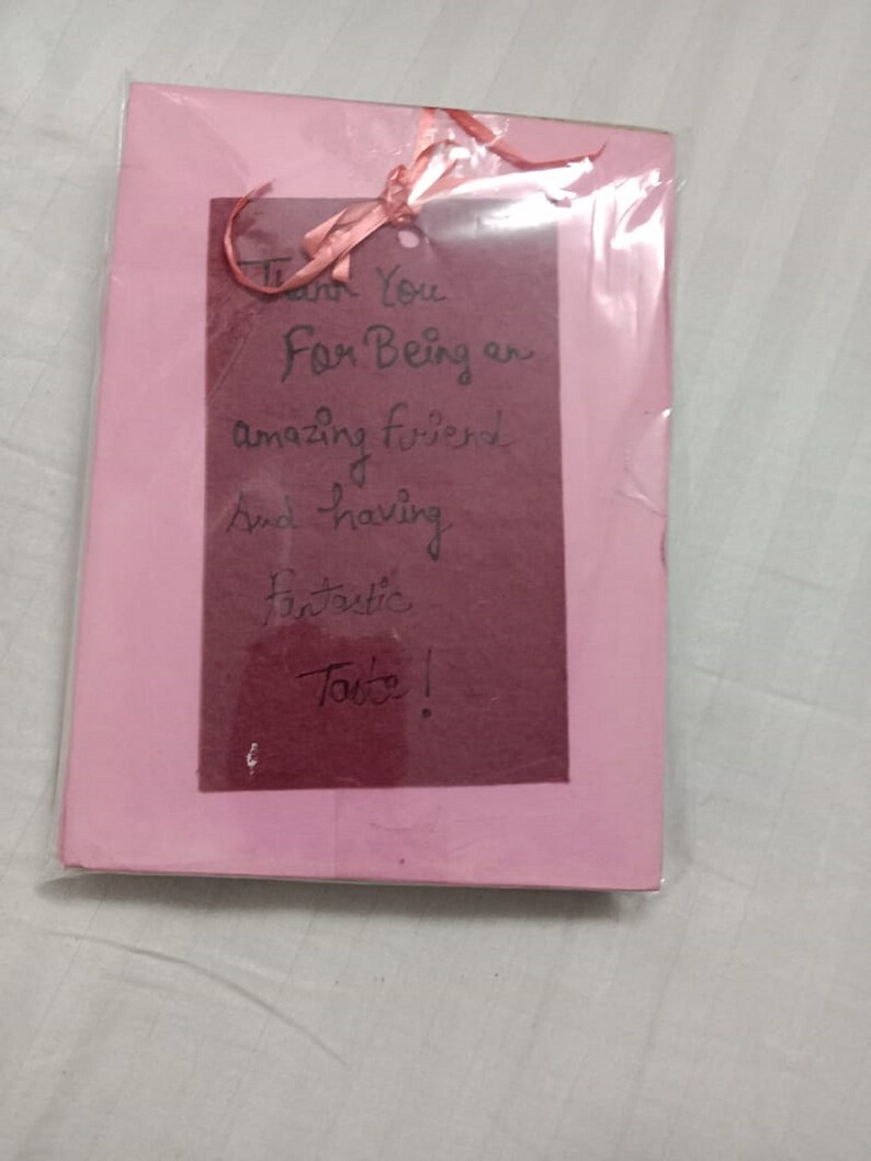Puede incluir: Una tarjeta de felicitaci&oacute;n rosa con un inserto rectangular rojo oscuro, envuelta en pl&aacute;stico transparente. Una cinta rosa est&aacute; atada en la parte superior. El texto manuscrito en el inserto dice: "Thank You For Being an amazing friend And having fantastic Taste!"