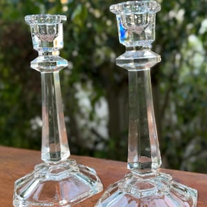 May include: Two clear glass candle holders with a faceted design. The candle holders have a hexagonal base and a tall, slender stem. The top of each holder has a small cup to hold a candle. The background is blurred green foliage.