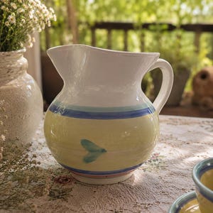 May include: A ceramic jug with a white top and a yellow body. The body features blue and pink stripes, and a teal leaf design. The jug has a handle and a spout. The jug is on a floral tablecloth.