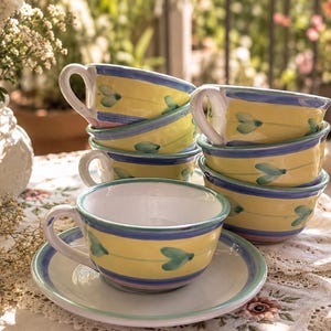 May include: A set of ceramic tea cups and saucers featuring a yellow base with horizontal blue and pink stripes and a repeating green heart-shaped leaf pattern. The cups have a white interior and a curved handle, with a matching saucer underneath.
