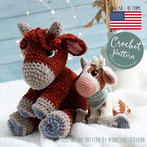 Op de afbeelding: Twee gehaakte koeienspeelgoed. De grotere koe is dieprood-bruin met grijze en blauwe accenten. De kleinere koe is wit met bruine vlekken, een roze neus en een bloemenkrans. De tekst "Crochet Pattern" is inbegrepen.
