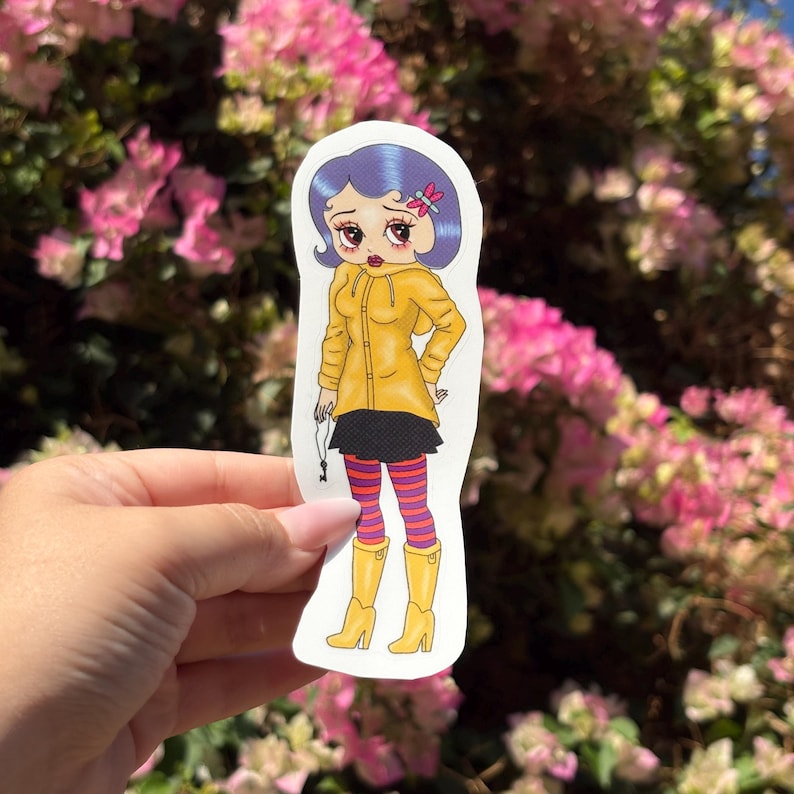 May include: A sticker featuring a cartoon illustration of a girl in a yellow raincoat, black skirt, and striped leggings. She has blue hair, a pink bow, and yellow boots. The girl is holding a key. The sticker is held in a hand against a background of pink flowers.