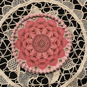 K&ouml;nnte beinhalten: Ein rosa Blumendesign, m&ouml;glicherweise ein Untersetzer oder Dekorationselement, ist auf einer schwarzen Oberfl&auml;che zentriert. Das Design zeigt eine detaillierte, geschichtete Blume mit einem Perlenmittelpunkt und ist von einem Ring aus kleinen rosa Pompons und Strasssteinen umgeben. Der Hintergrund ist eine cremefarbene Spitzendecke.