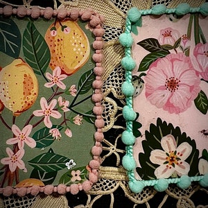 May include: Two square fabric panels with a decorative trim of pink and blue pom-poms. The panels feature floral and fruit patterns on a green and pink background.