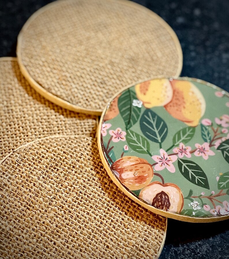 May include: Four round coasters, three are a natural burlap material and one is a green, floral design with peach and yellow fruit.