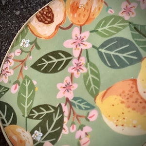 May include: A green ceramic plate with a floral pattern featuring pink flowers, green leaves, and yellow fruit. The plate has a white rim.
