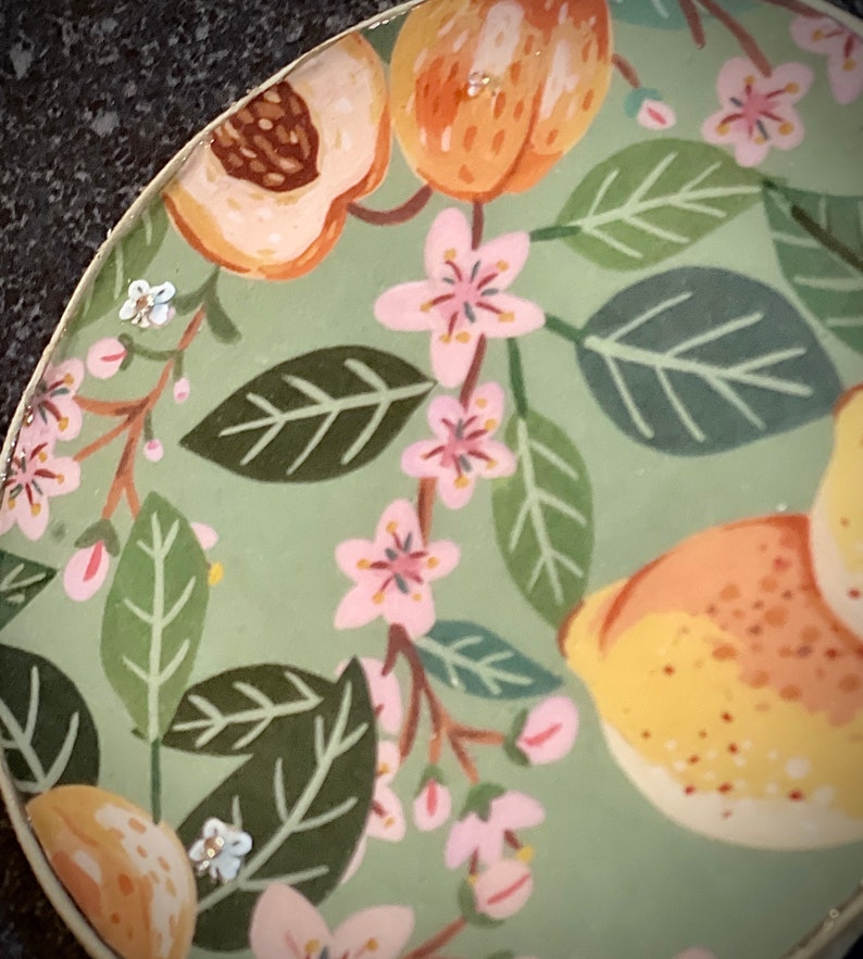 May include: A green ceramic plate with a floral pattern featuring pink flowers, green leaves, and yellow fruit. The plate has a white rim.